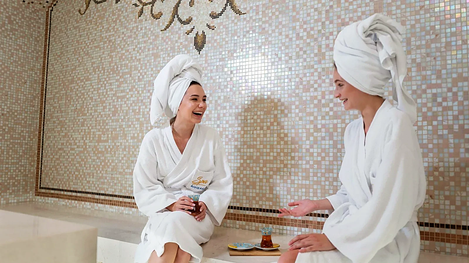 Two guests in bathrobes laughing over tea in a bright spa